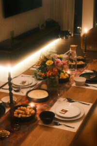 Friends gather around a warmly lit dining table with shared dishes, glasses, candles, and seasonal flowers.