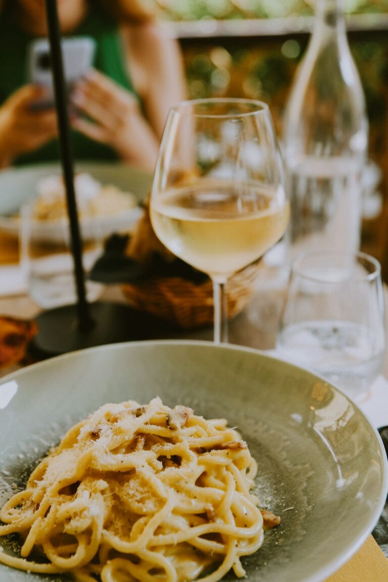 Several wine glasses sit beside steak, pasta, seafood, cheese, and vegetables on a warmly lit dining table.